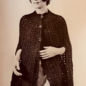 May include: A dark brown crocheted cape with three large buttons down the front. The cape has a scalloped hemline and a textured, open weave design. The image is a vintage photograph.