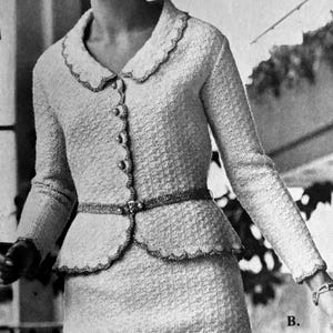 May include: A vintage-style, light-colored skirt suit. The jacket features a scalloped collar and hem, decorative buttons, and a matching belt. The skirt is knee-length. The suit has a textured weave.