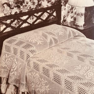 May include: A cream-colored, crocheted bedspread with a floral pattern, draped over a bed. The bed has a dark wooden headboard and a ruffled bed skirt. A floral patterned lamp sits on a nightstand.