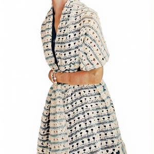 May include: A cream and light blue crocheted shawl featuring an openwork pattern with horizontal stripes and a fringe hem. The shawl is draped over a dark blue garment, showcasing a textured knit design with repeating triangular and linear motifs.