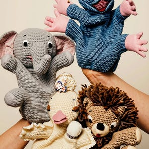 Op de afbeelding: Een set van vier gehaakte handpoppen, waaronder een blauwe kikker met roze handen, een grijze olifant met grote oren, een crèmekleurige eend met een kanten kraag en bloemaccessoire, en een beige leeuw met een donkerbruine manen.