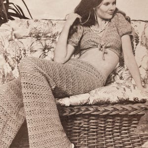 Peut inclure: Un ensemble deux pièces vintage au crochet de couleur marron clair. L'ensemble comprend un crop top à manches courtes avec une fermeture à nouer et un pantalon évasé. La tenue est portée par une personne assise sur un canapé à motifs floraux.