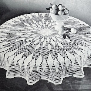 May include: A round, white crocheted tablecloth with a detailed geometric pattern. The design features a central circular motif with radiating lines and diamond shapes. A glass bowl with fruit sits on the table.