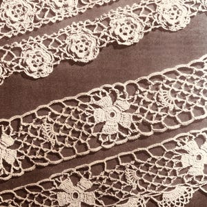 May include: Close-up of several strips of off-white crochet lace. The lace features floral and geometric patterns, with a scalloped edge on some strips. The intricate designs are visible against a dark background, highlighting the delicate craftsmanship.