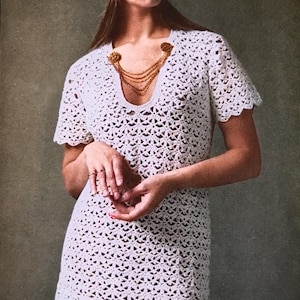 May include: A white crochet dress with short sleeves and a scalloped hem. The dress features a V-neckline adorned with a gold chain. The model is also wearing a white beret. The dress is a vintage style.