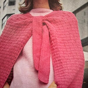 Puede incluir: Un chal de ganchillo rosa con un nudo atado en la parte delantera. El chal se lleva sobre un jersey rosa.