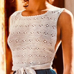 May include: A white, sleeveless crochet top with a scoop neckline and a tie-waist detail. The top is paired with a teal skirt. The crochet pattern features small, openwork designs. The overall style is vintage and handmade.
