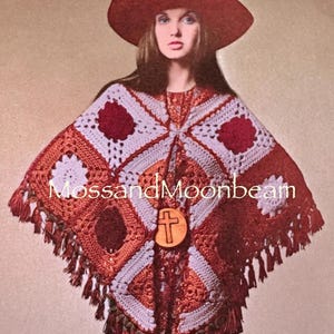 Könnte beinhalten: Ein gehäkelter Poncho in Rot-, Orange- und Weißtönen mit geometrischem Muster und Fransen. Ein Holzanhänger mit Kreuz hängt am Ausschnitt. Der Text "MossandMoonbeam" ist enthalten.