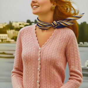 May include: A light pink long-sleeved cardigan with a V-neckline, a scalloped edge along the front placket, and five white round buttons. The knit fabric features a vertical textured pattern. A striped scarf in blue, yellow, and white is worn.