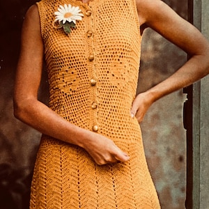 May include: A woman wearing a yellow crochet dress with a white daisy pinned to the front. The dress has a button-up front and a scalloped hem.