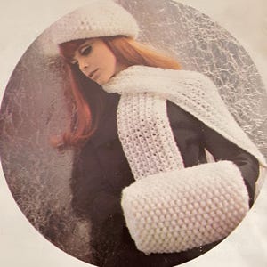 May include: A white knitted hat, scarf, and muff set. The scarf has an open weave design. The muff is a rectangular shape. The set is shown on a person with red hair and a black coat.
