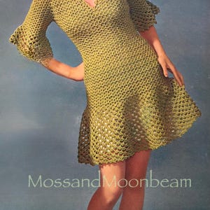 Op de afbeelding: Een vintage gehaakte jurk in een licht olijfgroene kleur. De jurk heeft een V-hals, korte mouwen met uitlopende manchetten en een uitlopende rok. De tekst "MossandMoonbeam" is zichtbaar onderaan de afbeelding.