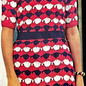 May include: A vintage-style dress with a red, white, and navy blue pattern. The dress features a fitted bodice, short sleeves, and a knee-length skirt. A dark blue belt cinches the waist. The woman is wearing a bracelet.