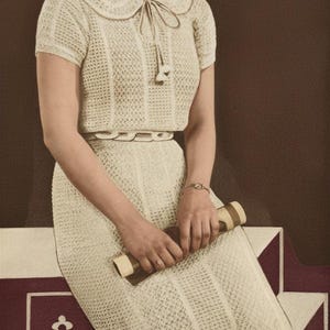 Op de afbeelding: Een crèmekleurige gehaakte jurk in vintage-stijl met een bijpassende top met korte mouwen en een kleine hoed. De jurk heeft een kraag met strik en een getailleerde taille. De vrouw houdt een opgerold voorwerp vast.