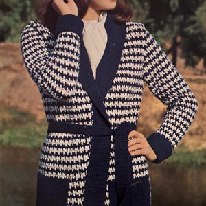 Könnte beinhalten: Eine langärmelige, knielange Strickjacke mit einem marineblau-weißen Hahnentrittmuster. Das Kleidungsstück verfügt über einen einfarbigen marineblauen Schalkragen, passende Bündchen, einen Bindegürtel und zwei Vordertaschen, kombiniert mit einem hellen Fischerhut.