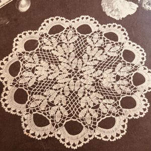May include: A white, circular doily with a detailed crochet pattern. The design features openwork circles around the edge and a central floral motif. The doily is made of a fine, delicate yarn.