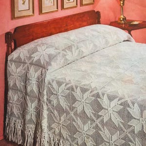 May include: A white, crocheted bedspread with a repeating starburst pattern. The bedspread has a fringed edge and is draped over a bed with a wooden headboard. The room has a pink wall and framed pictures.