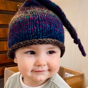 K&ouml;nnte beinhalten: Ein Baby tr&auml;gt eine bunte Strickm&uuml;tze mit einem langen Schwanz. Die M&uuml;tze hat einen braunen Rand und ist aus weichem, strukturiertem Garn gefertigt.