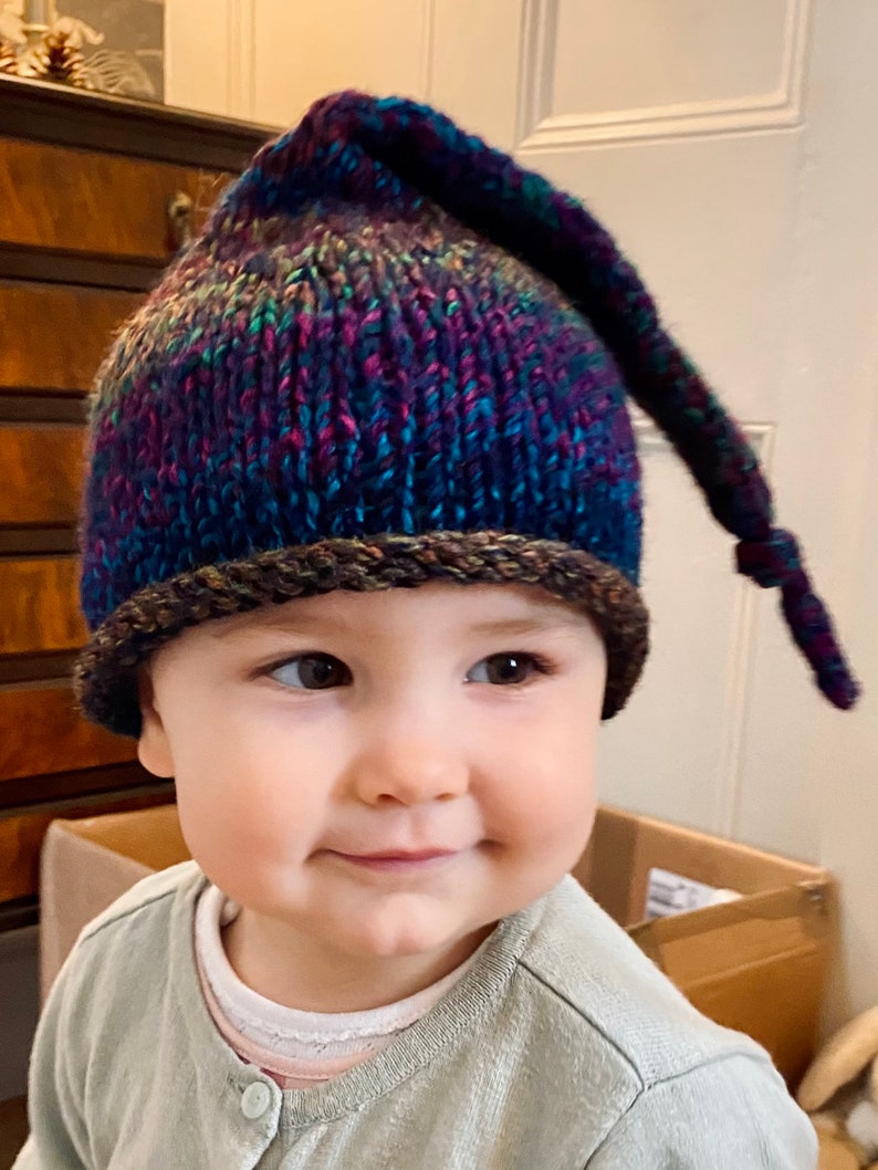 K&ouml;nnte beinhalten: Ein Baby tr&auml;gt eine bunte Strickm&uuml;tze mit einem langen Schwanz. Die M&uuml;tze hat einen braunen Rand und ist aus weichem, strukturiertem Garn gefertigt.