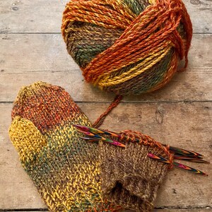 Peut inclure: Pelote de laine multicolore aux tons bruns, orang&eacute;s, jaunes et verts, &agrave; c&ocirc;t&eacute; d'une paire de mitaines tricot&eacute;es et d'aiguilles &agrave; tricoter. Les mitaines sont partiellement termin&eacute;es, r&eacute;v&eacute;lant la texture c&ocirc;tel&eacute;e du fil.
