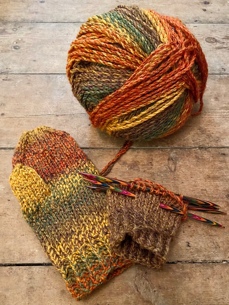 Peut inclure: Pelote de laine multicolore aux tons bruns, orang&eacute;s, jaunes et verts, &agrave; c&ocirc;t&eacute; d'une paire de mitaines tricot&eacute;es et d'aiguilles &agrave; tricoter. Les mitaines sont partiellement termin&eacute;es, r&eacute;v&eacute;lant la texture c&ocirc;tel&eacute;e du fil.
