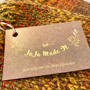 Peut inclure: Gros plan sur une &eacute;charpe tricot&eacute;e &agrave; la main dans des tons de jaune, orange et vert. Une &eacute;tiquette marron avec des lettres dor&eacute;es indique "JoJo Made It" et l'URL de la boutique Etsy. L'&eacute;charpe est en grosse laine.