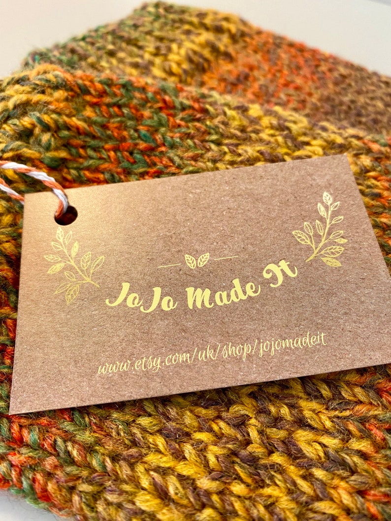 Peut inclure: Gros plan sur une &eacute;charpe tricot&eacute;e &agrave; la main dans des tons de jaune, orange et vert. Une &eacute;tiquette marron avec des lettres dor&eacute;es indique "JoJo Made It" et l'URL de la boutique Etsy. L'&eacute;charpe est en grosse laine.