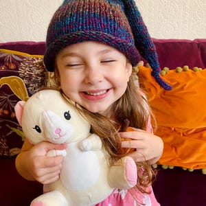 K&ouml;nnte beinhalten: Ein junges M&auml;dchen tr&auml;gt eine bunte Strickm&uuml;tze mit einer langen, spitzen Spitze und l&auml;chelt, w&auml;hrend es ein wei&szlig;es Pl&uuml;schtier mit rosa Akzenten h&auml;lt. Sie sitzt auf einem lila Sofa mit einem orangefarbenen Kissen hinter sich.