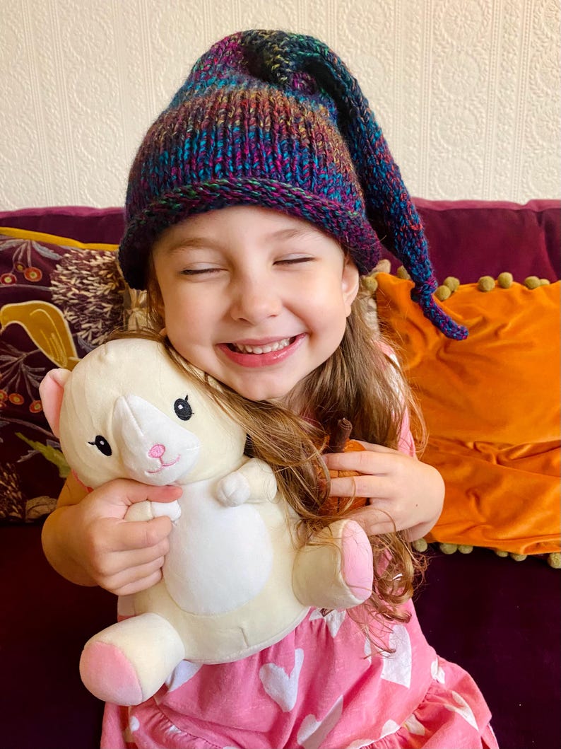 K&ouml;nnte beinhalten: Ein junges M&auml;dchen tr&auml;gt eine bunte Strickm&uuml;tze mit einer langen, spitzen Spitze und l&auml;chelt, w&auml;hrend es ein wei&szlig;es Pl&uuml;schtier mit rosa Akzenten h&auml;lt. Sie sitzt auf einem lila Sofa mit einem orangefarbenen Kissen hinter sich.