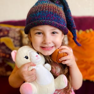 K&ouml;nnte beinhalten: Ein junges M&auml;dchen tr&auml;gt eine lila, blaue und gr&uuml;ne Strickm&uuml;tze mit einer langen Spitze und h&auml;lt ein wei&szlig;es Pl&uuml;schtier und einen braunen Basketball.
