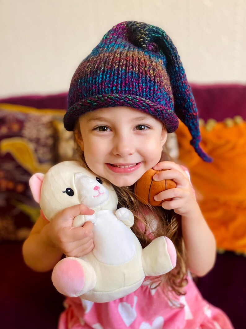 K&ouml;nnte beinhalten: Ein junges M&auml;dchen tr&auml;gt eine lila, blaue und gr&uuml;ne Strickm&uuml;tze mit einer langen Spitze und h&auml;lt ein wei&szlig;es Pl&uuml;schtier und einen braunen Basketball.