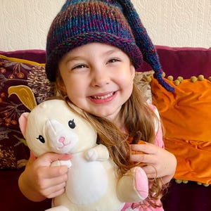 K&ouml;nnte beinhalten: Ein junges M&auml;dchen tr&auml;gt eine bunte Strickm&uuml;tze mit einer langen Spitze und h&auml;lt ein wei&szlig;es Pl&uuml;schkatzenspielzeug. Sie l&auml;chelt und schaut in die Kamera.