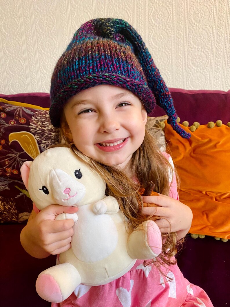 K&ouml;nnte beinhalten: Ein junges M&auml;dchen tr&auml;gt eine bunte Strickm&uuml;tze mit einer langen Spitze und h&auml;lt ein wei&szlig;es Pl&uuml;schkatzenspielzeug. Sie l&auml;chelt und schaut in die Kamera.
