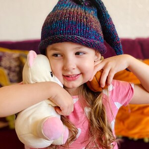 May include: A young girl wearing a purple, blue and green striped knitted hat with a long pointed tip, holding a white stuffed animal and a brown ball. She is wearing a pink shirt with white hearts.