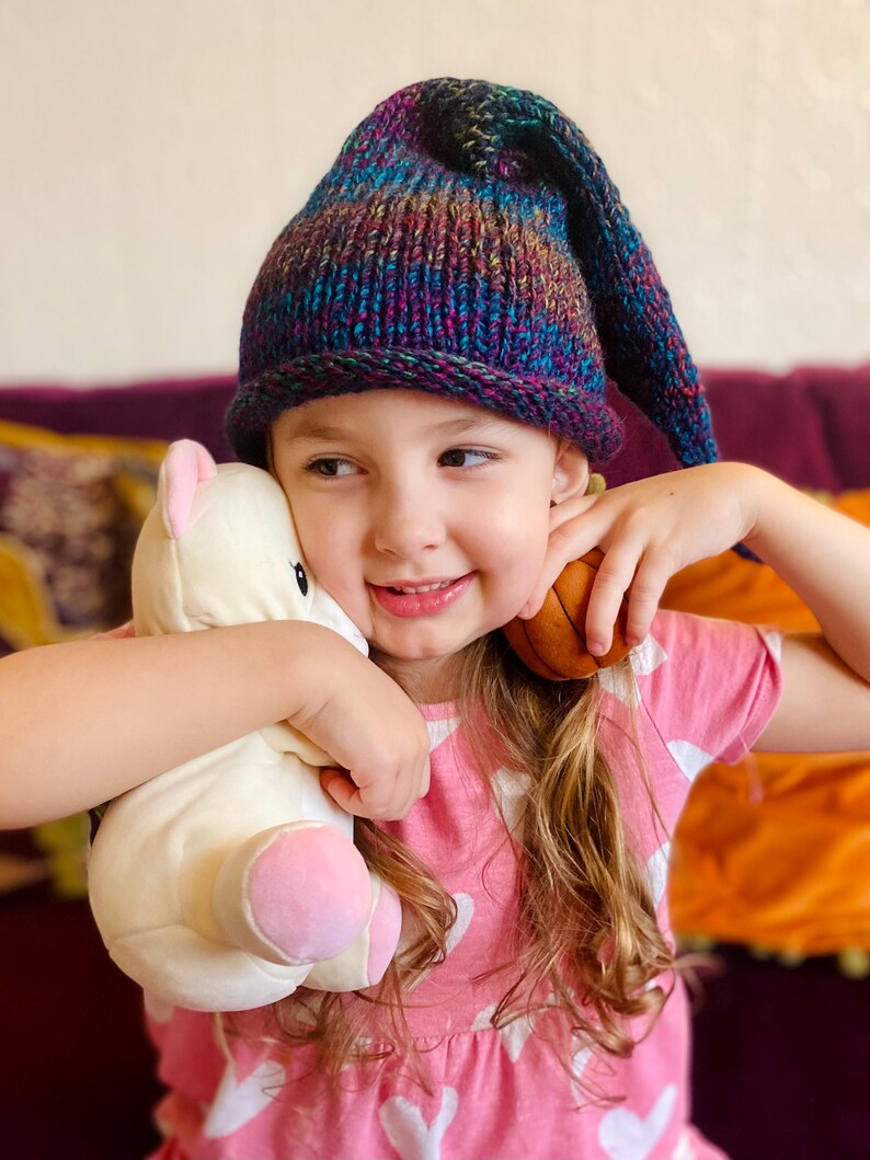 K&ouml;nnte beinhalten: Ein junges M&auml;dchen tr&auml;gt eine lila, blaue und gr&uuml;ne gestreifte Strickm&uuml;tze mit einer langen, spitzen Spitze, h&auml;lt ein wei&szlig;es Stofftier und einen braunen Ball. Sie tr&auml;gt ein rosa Shirt mit wei&szlig;en Herzen.