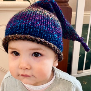 K&ouml;nnte beinhalten: Ein kleines Kind tr&auml;gt eine blaue, violette und braune Strickm&uuml;tze mit einer langen, spitzen Spitze. Die M&uuml;tze hat einen braunen Saum am unteren Rand.