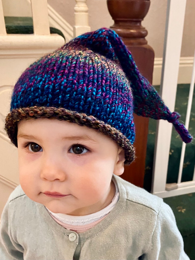 K&ouml;nnte beinhalten: Ein kleines Kind tr&auml;gt eine blaue, violette und braune Strickm&uuml;tze mit einer langen, spitzen Spitze. Die M&uuml;tze hat einen braunen Saum am unteren Rand.