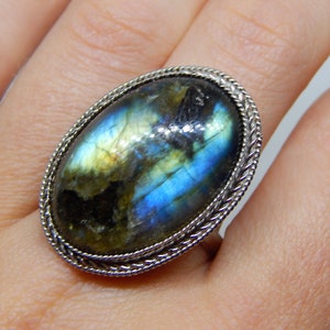 May include: A silver ring with a large oval-shaped labradorite gemstone. The stone has a blue, green, and gold iridescent sheen.