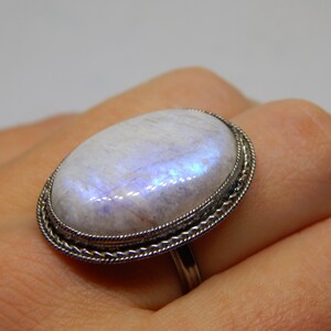 May include: A silver ring with a large oval moonstone set in a decorative silver bezel. The stone has a milky white color with iridescent blue and purple flashes.
