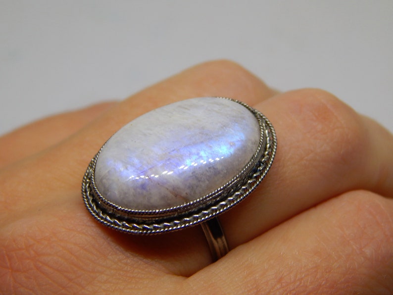 May include: A silver ring with a large oval moonstone set in a decorative silver bezel. The stone has a milky white color with iridescent blue and purple flashes.