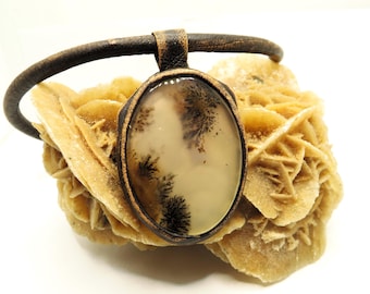 Puede incluir: Un collar de cordón de cuero marrón con un colgante que presenta una piedra de ágata blanca con inclusiones dendríticas negras. El colgante está engastado en un marco de metal de color bronce.
