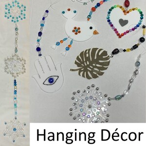 Puede incluir: Un móvil colgante colorido con varias formas y cuentas. Las formas incluyen un copo de nieve, una estrella, un pájaro, un búho, un corazón, una mano de hamsa y una hoja de monstera. Las cuentas son de varios colores, incluyendo azul, verde, rojo, naranja y morado. El móvil está etiquetado como "Decoración Colgante".