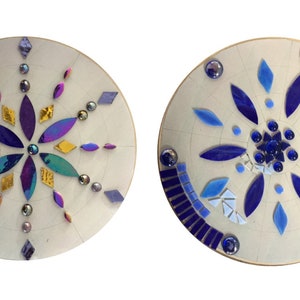 Puede incluir: Dos placas redondas de madera con dise&ntilde;os de mosaico. La primera placa tiene un dise&ntilde;o de flor morado, dorado y turquesa. La segunda placa tiene un dise&ntilde;o de flor azul.
