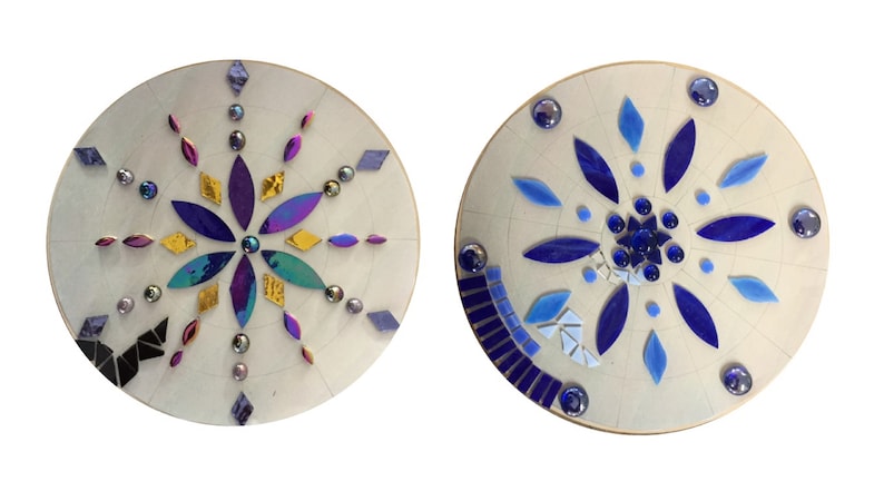 Puede incluir: Dos placas redondas de madera con dise&ntilde;os de mosaico. La primera placa tiene un dise&ntilde;o de flor morado, dorado y turquesa. La segunda placa tiene un dise&ntilde;o de flor azul.
