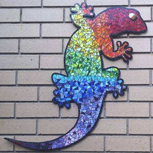Puede incluir: Un gecko de mosaico colorido con un patr&oacute;n de arco&iacute;ris. El gecko est&aacute; hecho de peque&ntilde;as baldosas de vidrio de colores y est&aacute; montado en una pared de ladrillo.