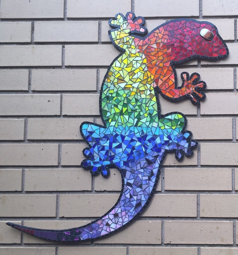 Puede incluir: Un gecko de mosaico colorido con un patr&oacute;n de arco&iacute;ris. El gecko est&aacute; hecho de peque&ntilde;as baldosas de vidrio de colores y est&aacute; montado en una pared de ladrillo.