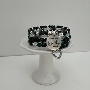 May include: A stack of beaded bracelets in shades of green, black, and clear, accented with silver beads. A silver charm with the word "Worthy" and a heart-shaped charm hangs from the bracelets. The bracelets are displayed on a white pedestal.
