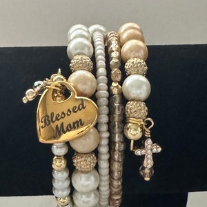 May include: A stack of beaded bracelets in shades of white, gold, and cream. One bracelet features a gold heart charm engraved with "Blessed Mom." Other charms include a cross. The bracelets are displayed on a black surface.