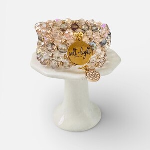 May include: Stacked beaded bracelets in shades of pink, gold, and clear crystals. A gold charm with the words "salt + light" is attached. The bracelets are displayed on a small white pedestal.