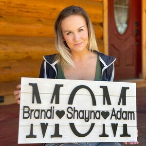 May include: A white wooden sign with the word "MOM" in large black letters. Below the word "MOM" are the names "Brandi", "Shayna", and "Adam" with a heart symbol between each name.
